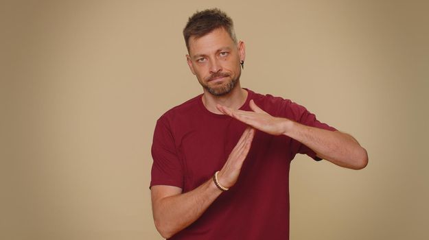 Time out, need a break. Tired serious upset handsome young man in red t-shirt showing pause relax gesture, limit or stop sign. Adult stylish male guy isolated alone on beige studio background indoors