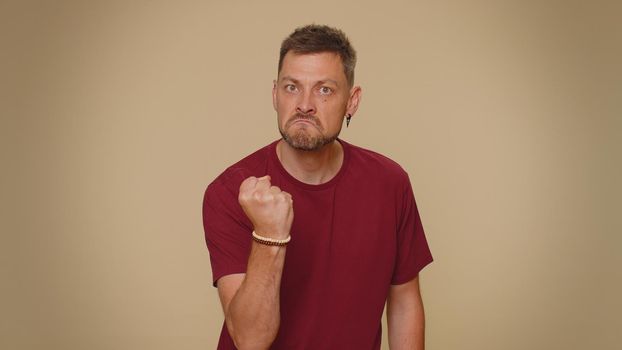 Aggressive angry handsome young man in red t-shirt trying to fight at camera, shaking fist, boxing with expression, punishment. Adult stylish male guy isolated alone on beige studio background indoors