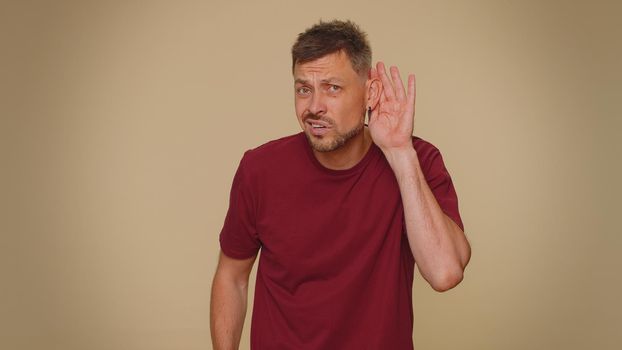 I cant hear you. What. Young man trying hear you, looking confused and frowning, keeping arm near ear for louder voice, asking to repeat, to hear information, deafness. One guy on beige background