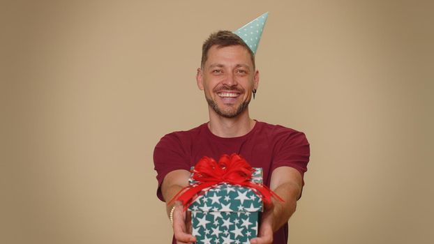 Positive smiling bearded handsome young man presenting birthday gift box stretches out hands, offer wrapped present career bonus, celebrating party. Adult stylish guy on beige studio background indoor