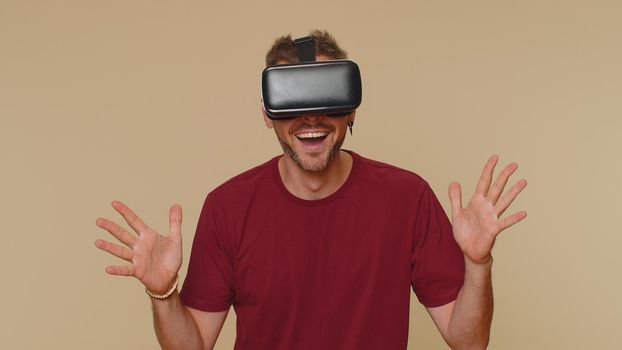 Bearded handsome young man using headset helmet app to play simulation drawing game. Watching virtual reality 3D 360 video. Adult male guy in VR goggles isolated on beige studio background indoors