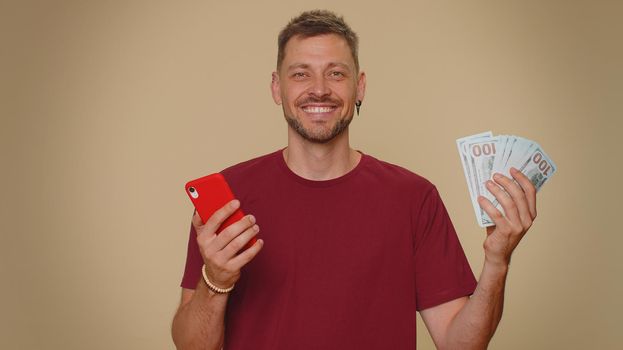 Happy young man in red t-shirt looking smartphone display sincerely rejoicing win, receiving money dollar cash banknotes, success lottery luck. Adult stylish male guy on beige studio background