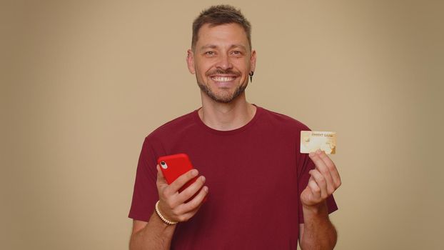 Happy young man use smartphone, plastic credit bank card, browsing say wow yes, found out great big win, success lotter, good news, purchases online shopping. Adult guy on beige studio background