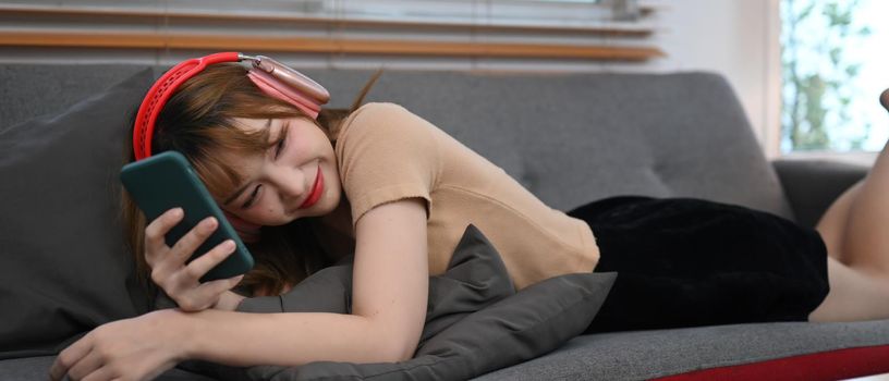 Cheerful young woman resting on couch and using mobile phone, spending time in social media internet.