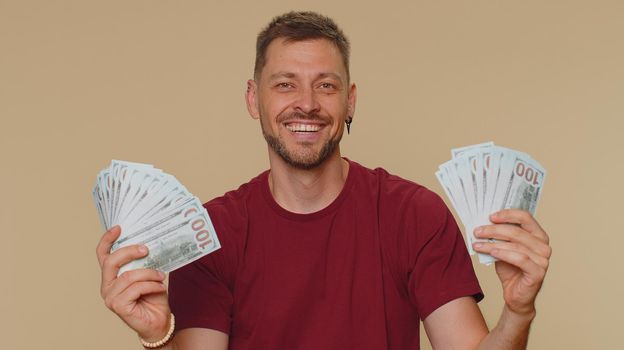 Bearded handsome young man holding fan of cash money dollar banknotes celebrate dance, success business career, lottery game winner, big income, wealth. Adult stylish guy on beige studio background