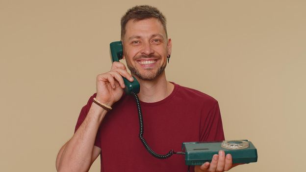 Hey you, call me back. Handsome young man in red t-shirt talking on wired vintage telephone of 80s, says hey you call me back. Call center. Adult stylish male guy on beige studio background indoor