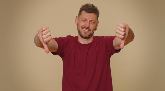 Upset bearded handsome young man showing thumbs down sign gesture, expressing discontent, disapproval, dissatisfied, dislike. Adult stylish male guy isolated alone on beige studio background indoors