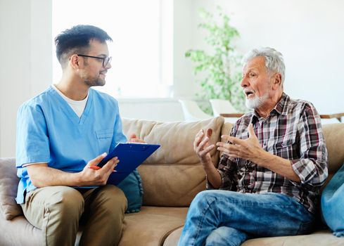 Doctor or nurse caregiver talking with senior man on sofa at home or nursing home