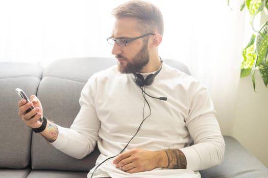 Glad fashionable young bearded guy wears stylish clothes and denim shirt, has trendy hairstyle, happy to exchange messages with friends, uses free internet connection on electronic modern gadget