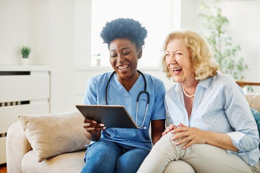 Doctor or nurse caregiver showing a tablet screen to senior woman and laughing at home or nursing home