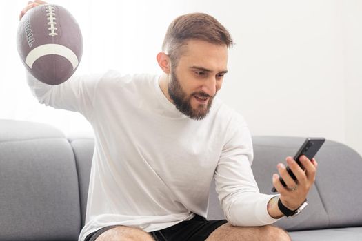 American football fan holding rugby ball and watching match on smartphone.