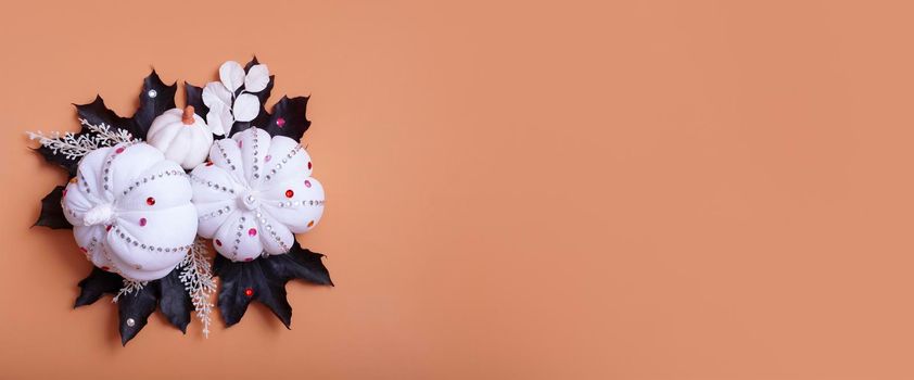 Creative autumn banner with flat lay composition from white textile decorative pumpkins and dark maple leaves decorated rhinestones on orange background. Copy space.