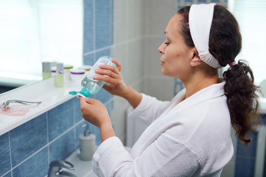 Side portrait of a beautiful curly-haired brunette woman in a white bathrobe, pouring a cleansing lotion on a cotton pad to remove makeup from her skin, standing in front of a mirror in the bathroom