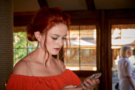 an adult female human being. Serious cute redhead young woman in red dress reading a message in the phone.