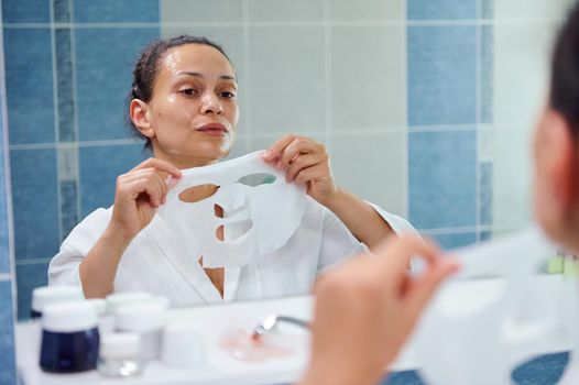 Reflection in bathroom mirror of a beautiful dark-haired multi-ethnic woman, in white bathrobe applying a smoothing moisturizing textile face mask. Skin care. Home spa. Anti-aging beauty procedures