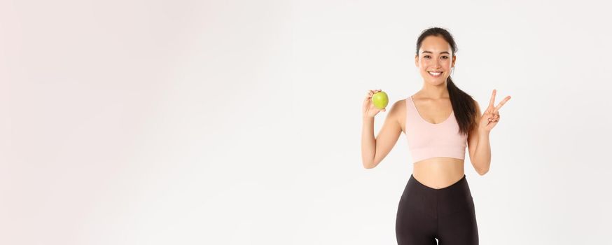 Sport, wellbeing and active lifestyle concept. Attractive asian female fitness coach, girl trainer advice eating healthy food after workout and training, standing with apple, show peace sign.
