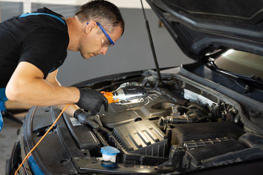 Professional Mechanic in Blue Overalls is Working on a Car in a Car Service. Repairman in Safety Glasses is Working on an Usual Car Maintenance. He Hangs a Lamp. Modern Workshop.