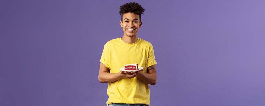 Celebration, party and holidays concept. Portrait of handsome young smiling man, feeling happy likes eating desserts, ordered delivery from local cafe to have some delicious cake, purple background.