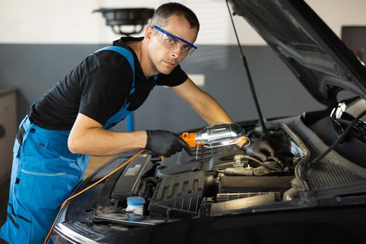 Portrait of young caucasian man in blue overalls and safety glasses inspects engine with flashlight. Male car mechanic at work in spacious repair shop. Modern workshop.