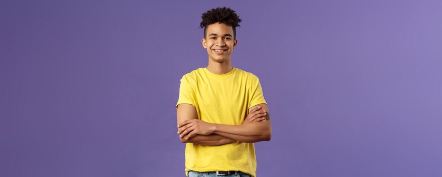 Close-up portrait of confident, smart and professional young male student with dreads, yellow t-shirt, cross arms over chest and smiling pleased, knows what he doing, purple background.