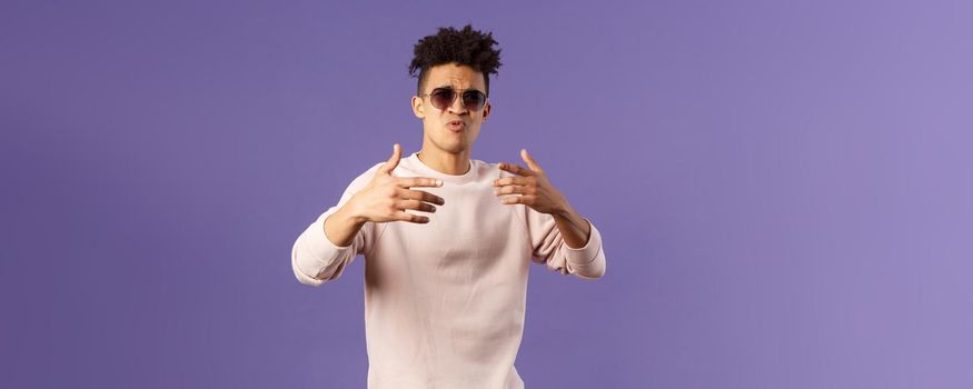 Portrait of sassy and carefree young hipster guy with dreads, wearing sunglasses dancing and gesturing while singing during rap battle, dance hip-hop on party, standing purple background.