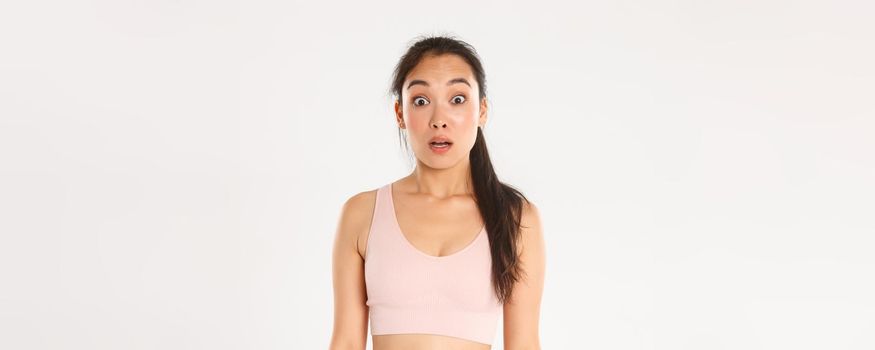Sport, wellbeing and active lifestyle concept. Close-up of surprised and startled asian fitness girl, sportswoman in sportsbra looking confused with dropped jaw, hear news, white background.