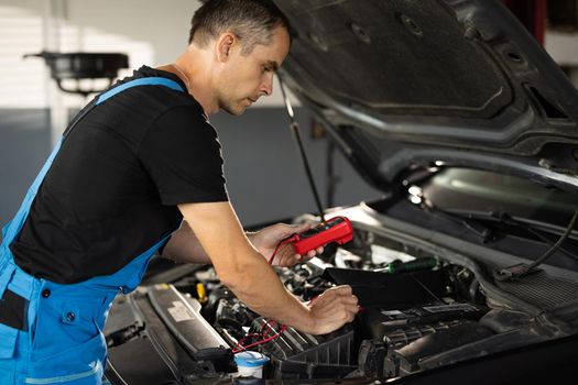 Professional car mechanic check battery voltage with electric multimeter. Automobile diagnosis. Car mechanic repairer looks for engine failure on diagnostics equipment in vehicle service workshop.