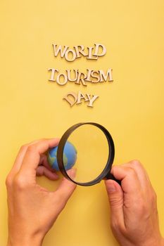 World Tourism Day text from wooden letters on a yellow color background with globe in female hands and a magnifying glass.