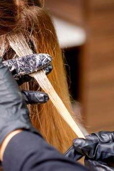 Closeup back view of hairdresser's hands in gloves applying dye to a strand of hair of redhead young woman in a hair salon