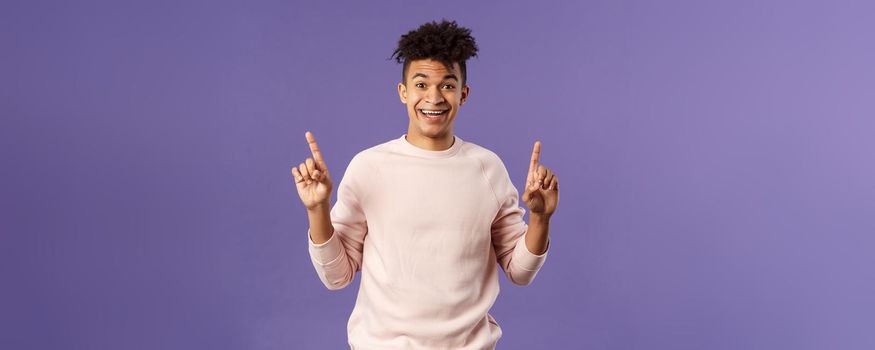 Portrait of charismatic lively young man heard about good deal, asking question intrigued, pointing fingers up, smiling upbeat at camera, look with interest, recommend product, purple background.