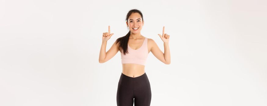 Sport, wellbeing and active lifestyle concept. Cheerful good-looking asian sportswoman in sportsbra and leggings pointing and looking up, smiling pleased, found good promo offer.