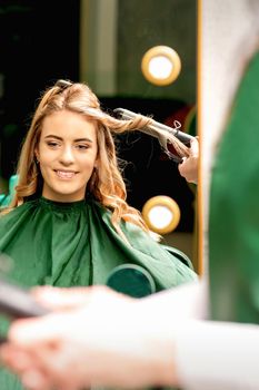 Hairdresser using curling tongs curls long brown hair on the young caucasian girl in a beauty salon