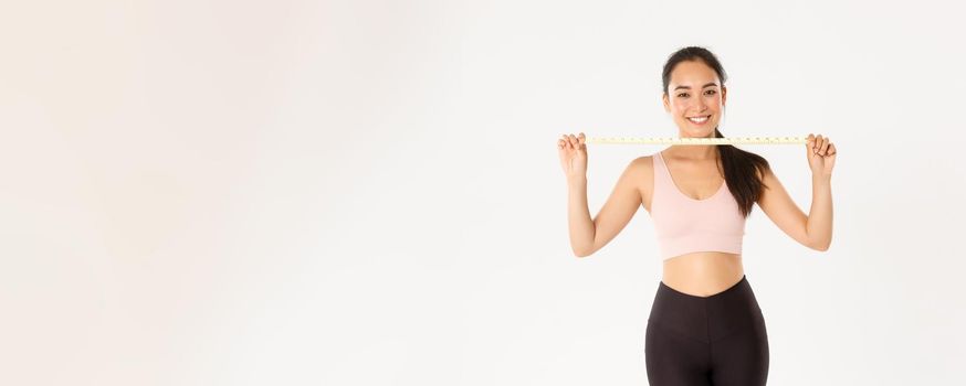 Fitness, healthy lifestyle and wellbeing concept. Portrait of smiling asian brunette sports girl, female athelte in activewear showing measuring tape, losing weight with exercises, white background.