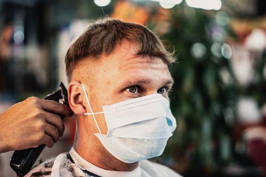 Lifestyle photo. Professional barbershop. Young man customer wears face mask. Hairdresser male hands work move comb tool electric clipper. Modern style short hair locks cut. Fashion service salon.