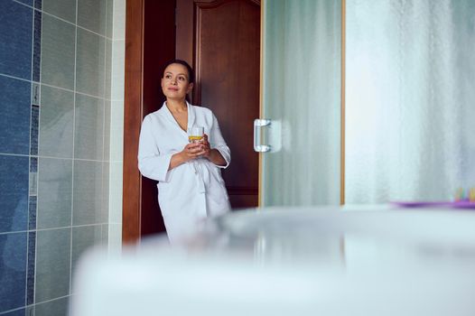 Gorgeous confident cute multi-ethnic middle-aged woman wearing a white bathrobe, standing in a cozy home bathroom with a glass of lemon water in her hands. Diet. Vitality. Healthy lifestyle. Body care