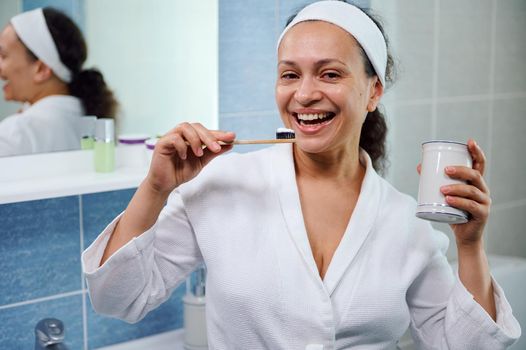 Multiethnic middle aged woman in white bathrobe, enjoys morning beauty procedures, brushing teeth with eco bamboo toothbrush and whitening toothpaste, smiles beautiful toothy smile, looking at camera