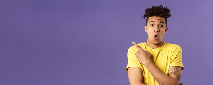 Portrait of surprised, excited young man showing cool new thing, pointing upper left corner, gasping, folding lips, stare camera impressed say wow, standing purple background.