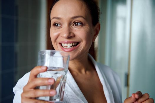 Close-up portrait of a beautiful happy multi-ethnic middle-aged woman smiling, taking medication with nutritional dietary supplements, according to physician's prescription and drinking mineral water