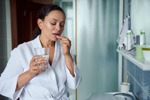 Middle-aged multi-ethnic woman in white bathrobe, taking daily dose of complex healthcare skin, hair and nails omega vitamins and drinking glass of fresh pure water. Immunity improvement concept.