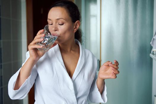Attractive young dark-haired woman in white bathrobe, holding pill and glass of fresh pure water, taking antioxidant medicine vitamins, beauty supplements for hair skin nails. Healthcare concept.