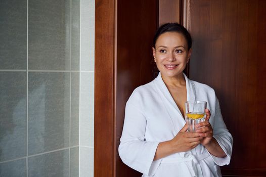 Close-up portrait of a stunning multi-ethnic brunette woman with glowing clean and healthy skin, in bathrobe, holding glass with lemon water for good start to the day. Diet, vitality, healthy habits
