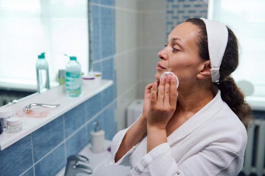 Close-up portrait of a natural beauty woman in white bathrobe, using cleanser cosmetic lotion and cotton pad, removes makeup, enjoys skin care procedures, looking at her reflection in bathroom mirror