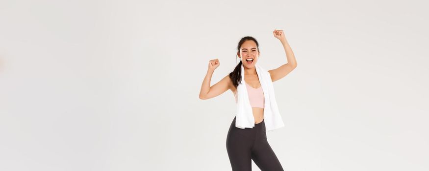 Sport, gym and healthy body concept. Full length of happy asian brunette girl, slim female athelte in active wear, shouting yes and raising hands up motivated to workout, enjoy training in gym.