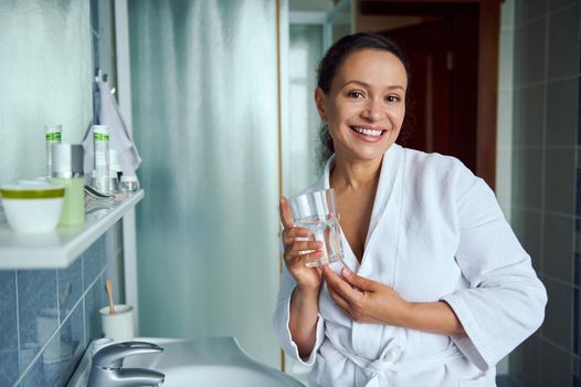 Charming stunning attractive multi-ethnic woman in white bathrobe, smiling a cheerful toothy smile, standing with a glass of mineral water in the bathroom. Morning routine. Healthy lifestyle