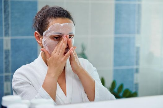 Reflection in the bathroom mirror of multi-ethnic middle-aged woman, massaging her face while applying smoothing moisturizing sheet mask on her face. Home spa, beauty treatment for glowing clean skin