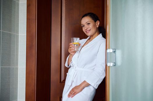 Diet, vitality, beauty care and healthy lifestyle, morning routine. Calm cute African American woman wearing white waffle bathrobe, drinking clean water with lemon at glass in cozy home bathroom