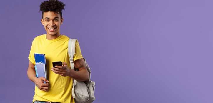 Back to school, university concept. Portrait of young handsome smiling man, student asking for classmate phone number, making note in mobile phone, wear backpack, hold notebooks and study material.