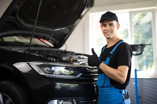 Portrait of car mechanic in a car workshop shows thumbs up, in the background of service. Positive auto service worker smiling to camera and showing thumb up gesture, approving car repair workshop
