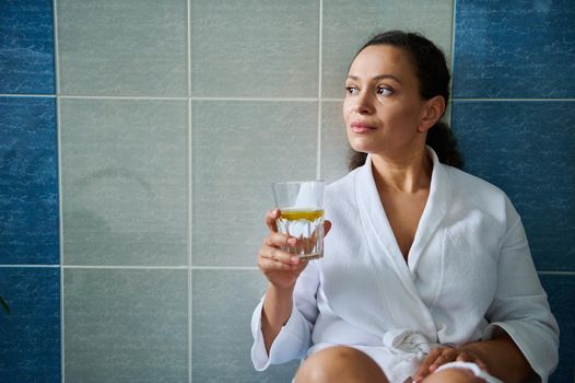 Multi-ethnic delightful middle-aged pretty woman, in a white bathroom, holding a glass of lemon water, taking care of her health. Healthy habits. Diet Vitality. Good start to the day. Morning routine