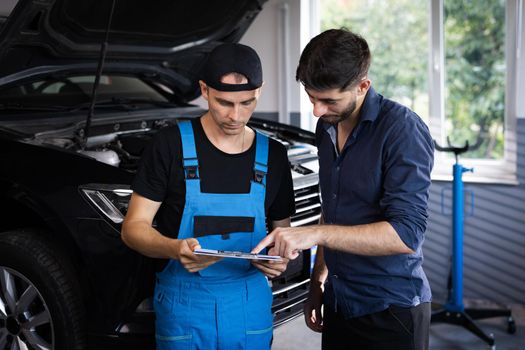 Car service manager with a tablet, talking to mechanic man discussing car diagnostics and repairing. Mechanic talking to manager near vehicle in car service. Specialist is showing info on tablet